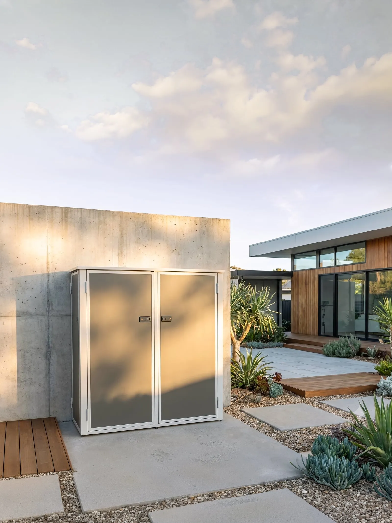 Garden S storage unit with grey doors against a concrete wall in a courtyard garden at dusk